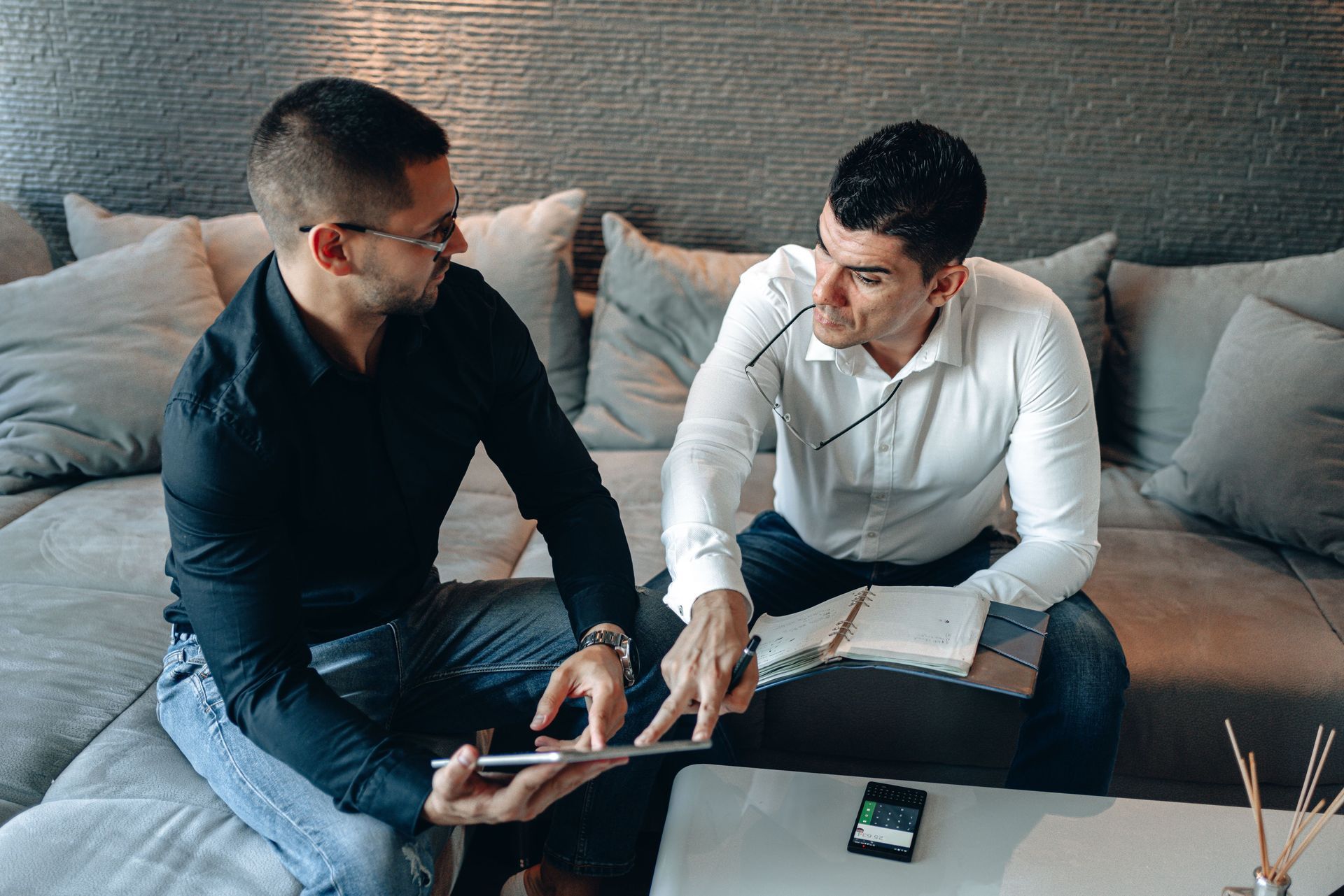 Two people talking over a tablet on a couch in a living room, with a smartphone on the table.