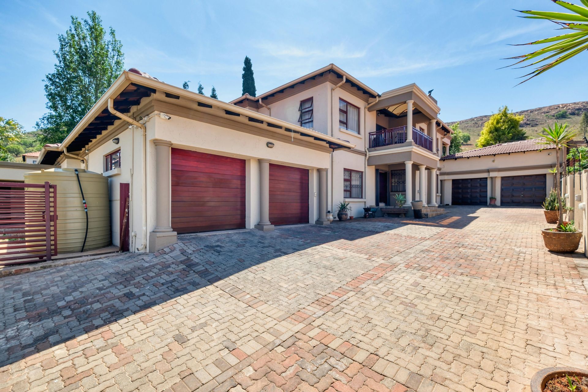 Two-story house with brick driveway, three red garage doors, and balcony, in a sunny setting.