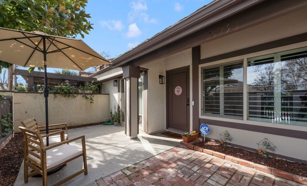 A patio area features a tan umbrella, two wooden chairs, a brick border, and a home entrance with a large window.