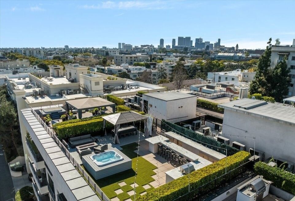 Rooftop terrace with a hot tub, lounge area, and outdoor kitchen, overlooking a city skyline on a sunny day.