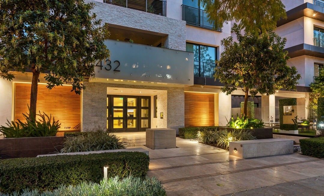 Modern building entrance with numbered address 432, lit by warm lights at dusk, surrounded by manicured trees and shrubs.