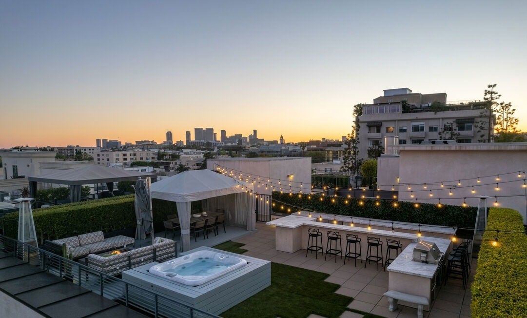 A rooftop terrace at sunset, featuring a hot tub, lounge area, and bar with string lights overlooking a city skyline.