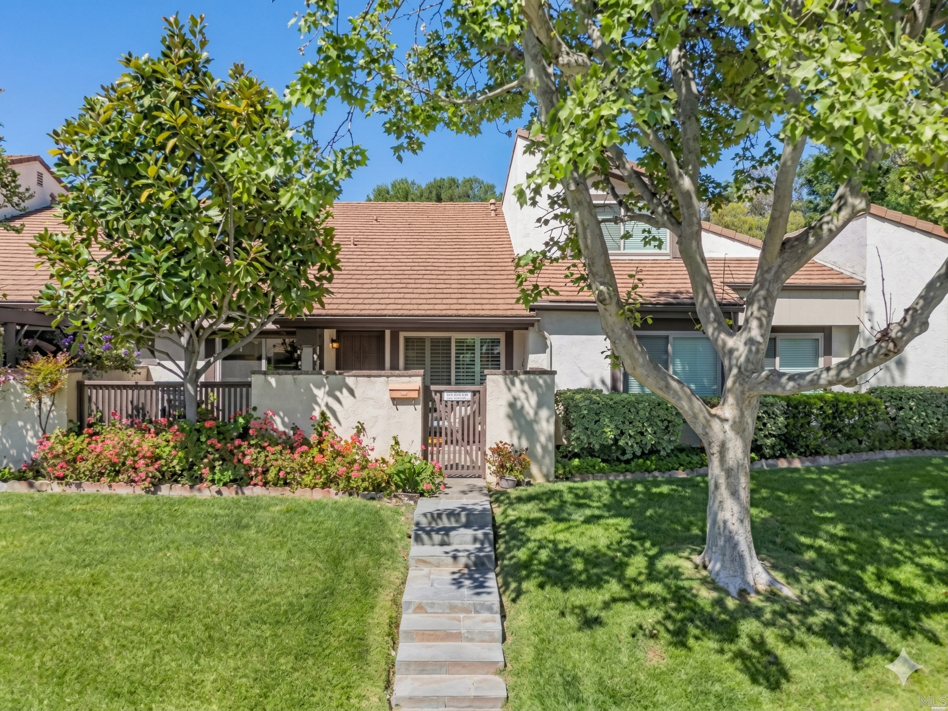 A tan townhome with a brown shingled roof, a stone walkway, green lawn, and trees under a clear blue sky.
