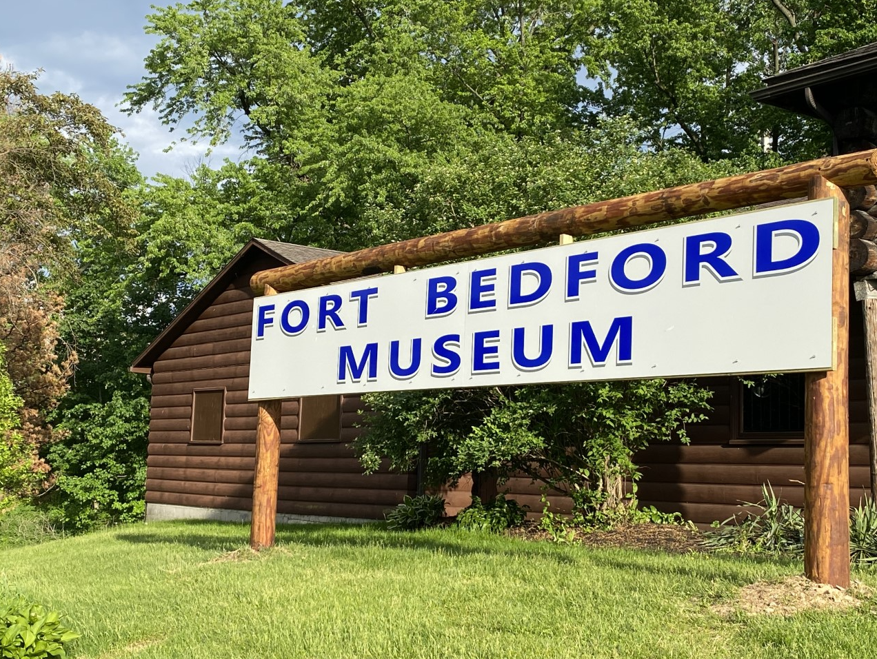 A sign for the fort bedford museum is in front of a log cabin.