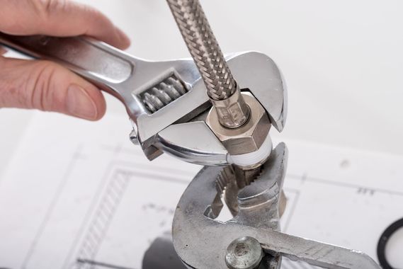 A hand uses an adjustable wrench and pliers to tighten a metal hose fitting over a technical blueprint.