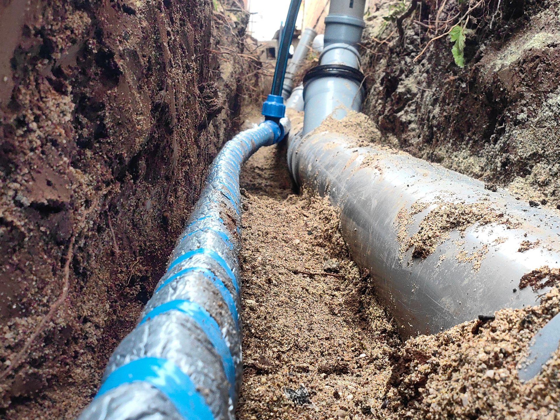 A trench shows a blue polyethylene water pipe running alongside a larger grey PVC drainage pipe in a sandy bed. A trench shows a blue polyethylene water pipe running alongside a larger grey PVC drainage pipe in a sandy bed.