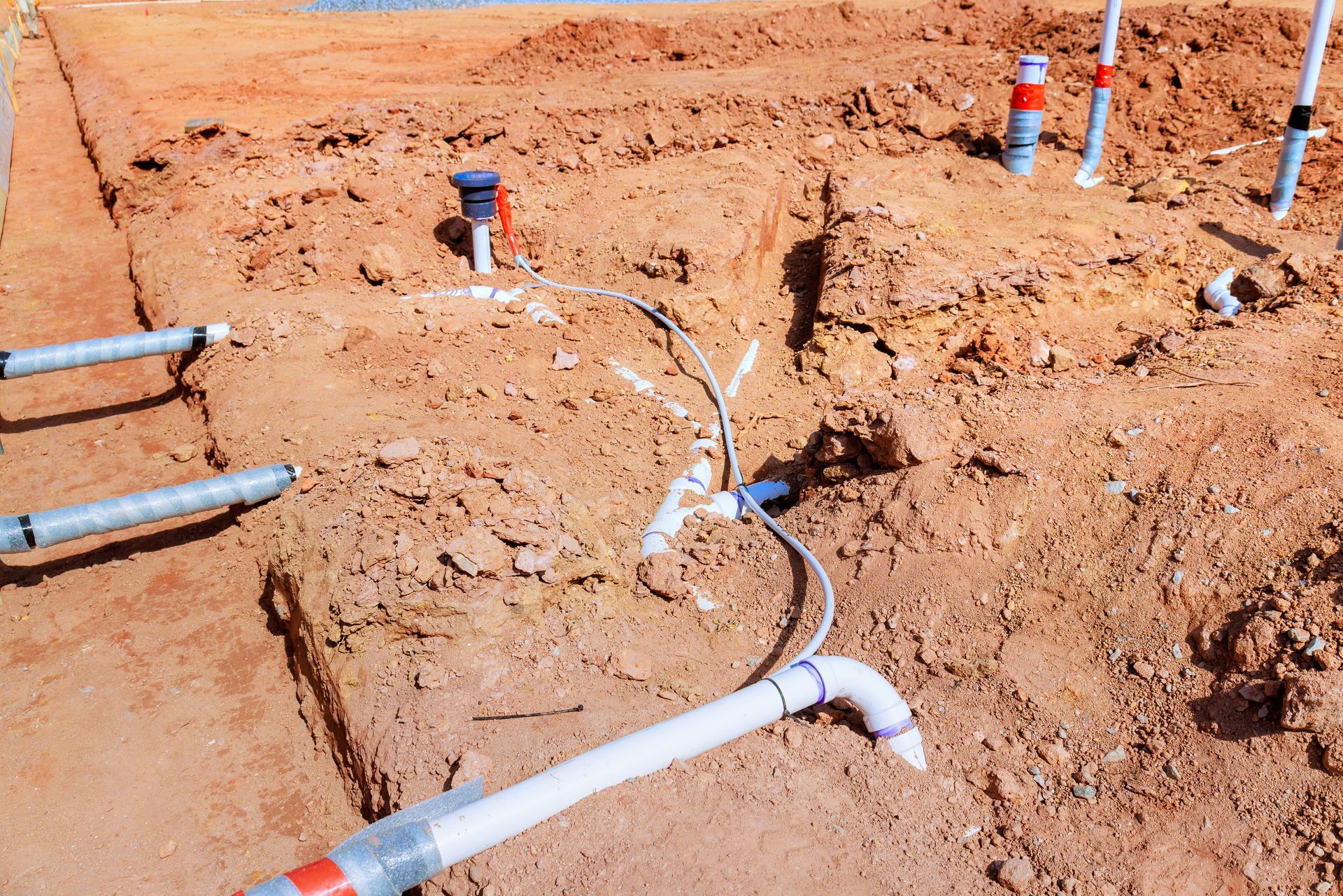 Construction site showing white PVC pipes, electrical conduits, and wiring laid out in dirt trenches for slab foundation. Construction site showing white PVC pipes, electrical conduits, and wiring laid out in dirt trenches for slab foundation.