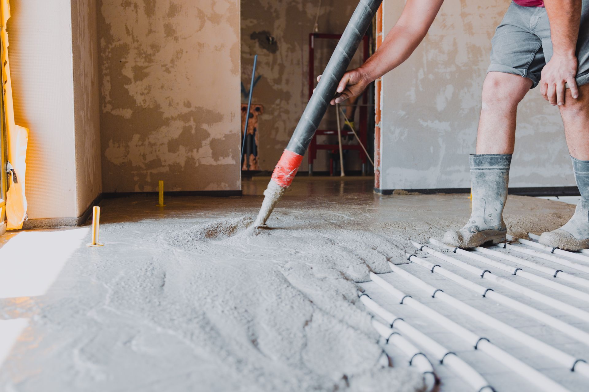 A worker in boots uses a hose to pour liquid concrete over radiant floor heating pipes in a room under construction. A worker in boots uses a hose to pour liquid concrete over radiant floor heating pipes in a room under construction.