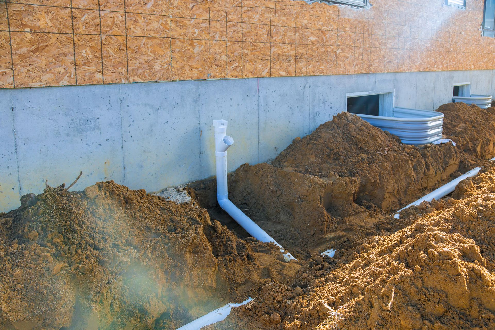 Construction site showing a house foundation with exposed white plumbing pipes and window wells surrounded by piles of dirt.