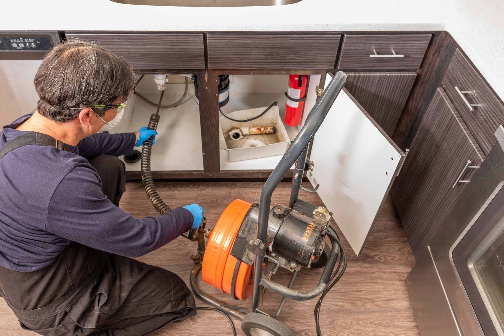 A plumber in blue gloves uses a professional orange drain auger to clear a clog under a kitchen sink. A plumber in blue gloves uses a professional orange drain auger to clear a clog under a kitchen sink.