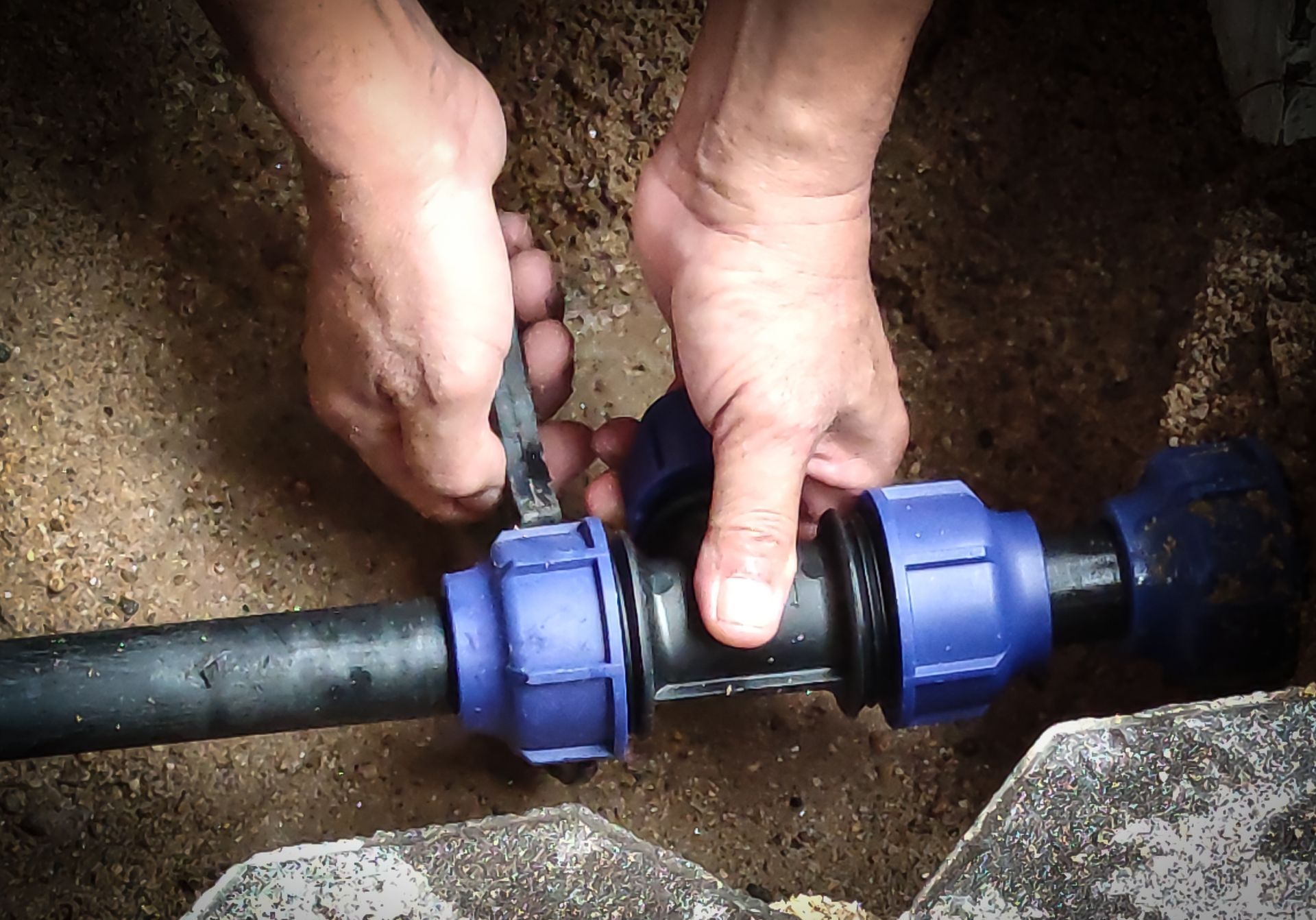 A person’s hands tighten a blue compression coupling onto a black pipe in a dirt trench. A person’s hands tighten a blue compression coupling onto a black pipe in a dirt trench.