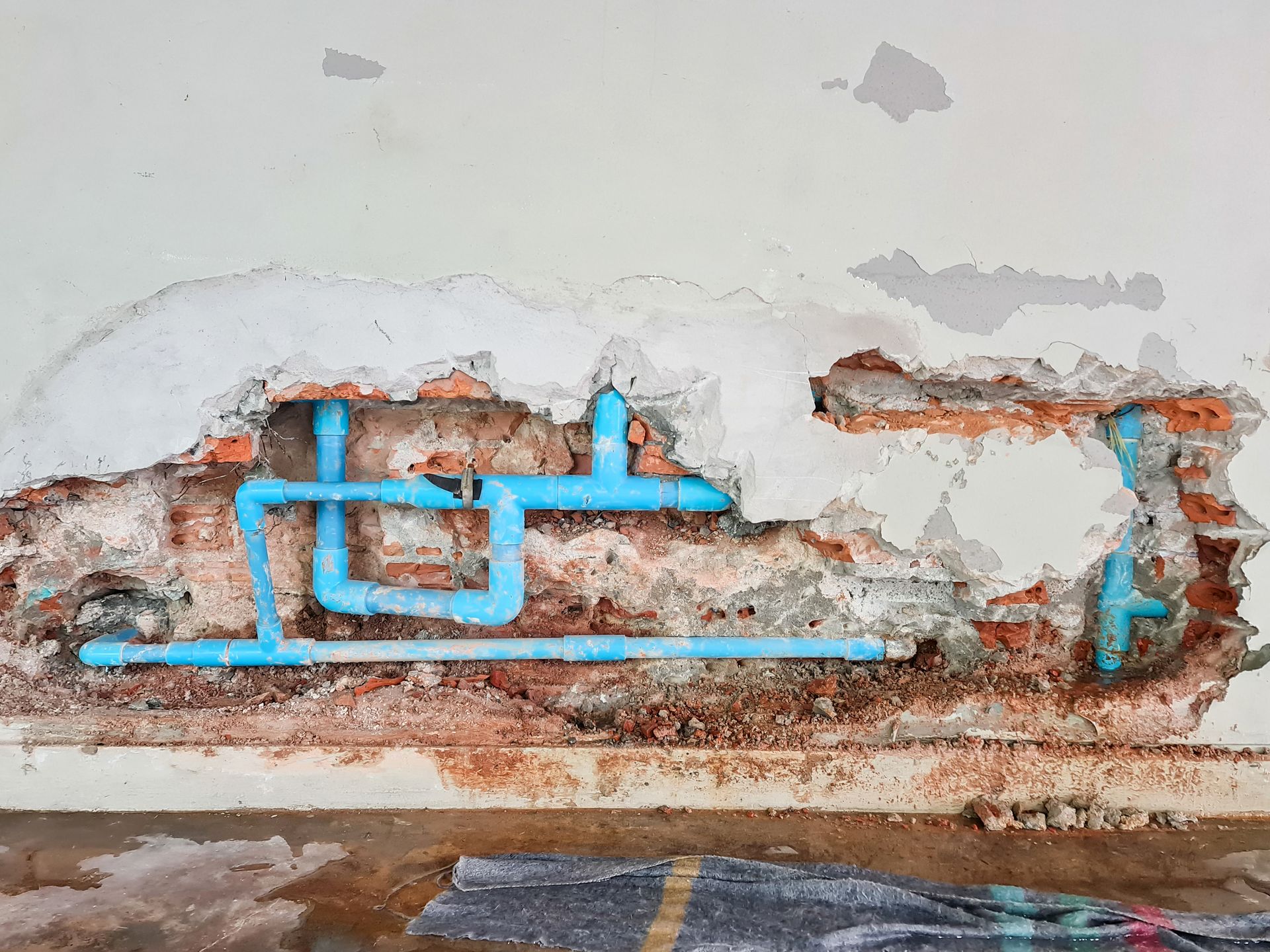 Exposed blue PVC plumbing pipes running through a section of broken, unfinished brick wall with crumbling plaster.