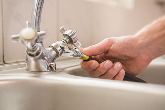 A hand uses an adjustable wrench to tighten a metal faucet over a kitchen sink.