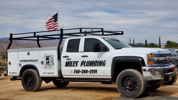 White Miles Plumbing utility truck with a ladder rack and American flag parked in an outdoor, dirt lot.