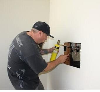 A worker uses a yellow handheld torch to solder copper pipes inside an open section of a white wall. A worker uses a yellow handheld torch to solder copper pipes inside an open section of a white wall.