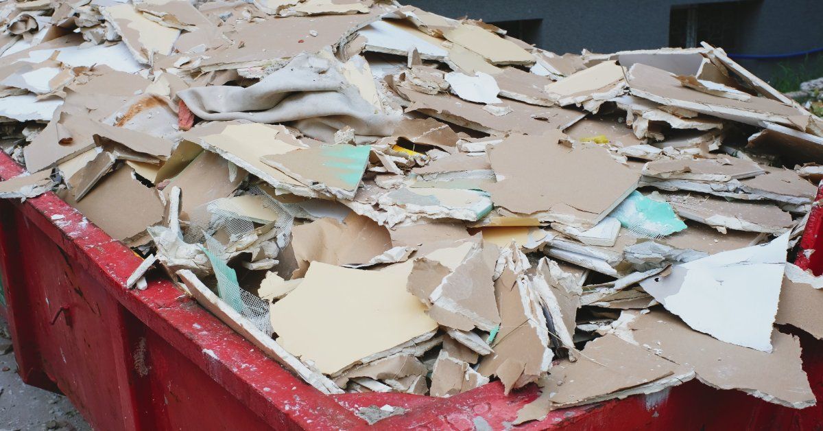 A large metal dumpster filled with broken construction debris, white bags, wood pieces, and loose ma
