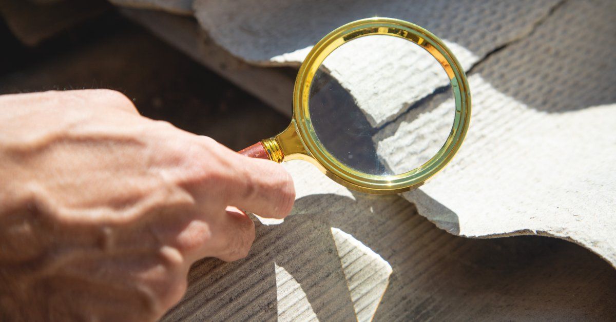 A person holding a magnifying glass over corrugated roofing material with broken edges