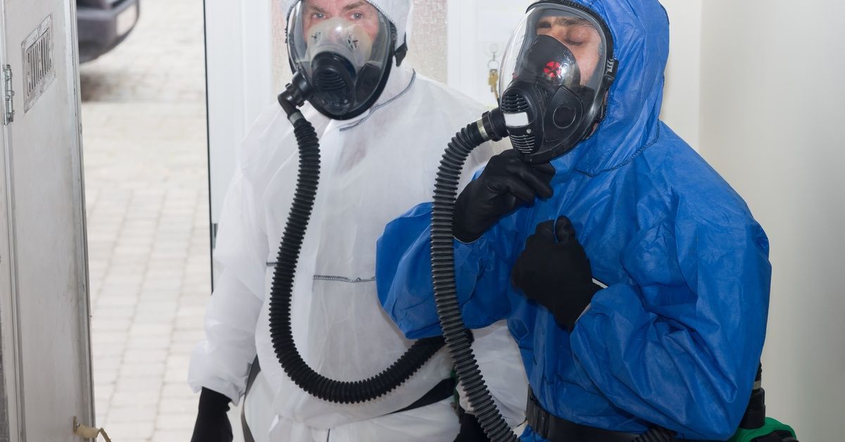 Two men in coveralls and respirator masks adjust their protective gear as they stand by the doorway