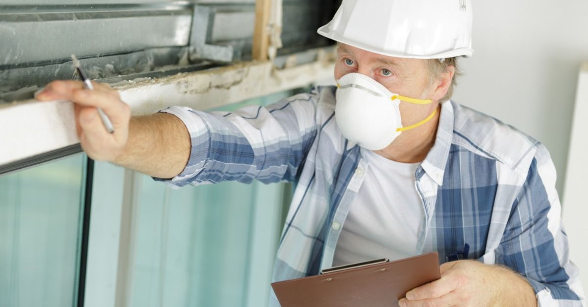 A close-up of a man wearing a face mask and gloves, inspecting a wall with peeling paint and damage