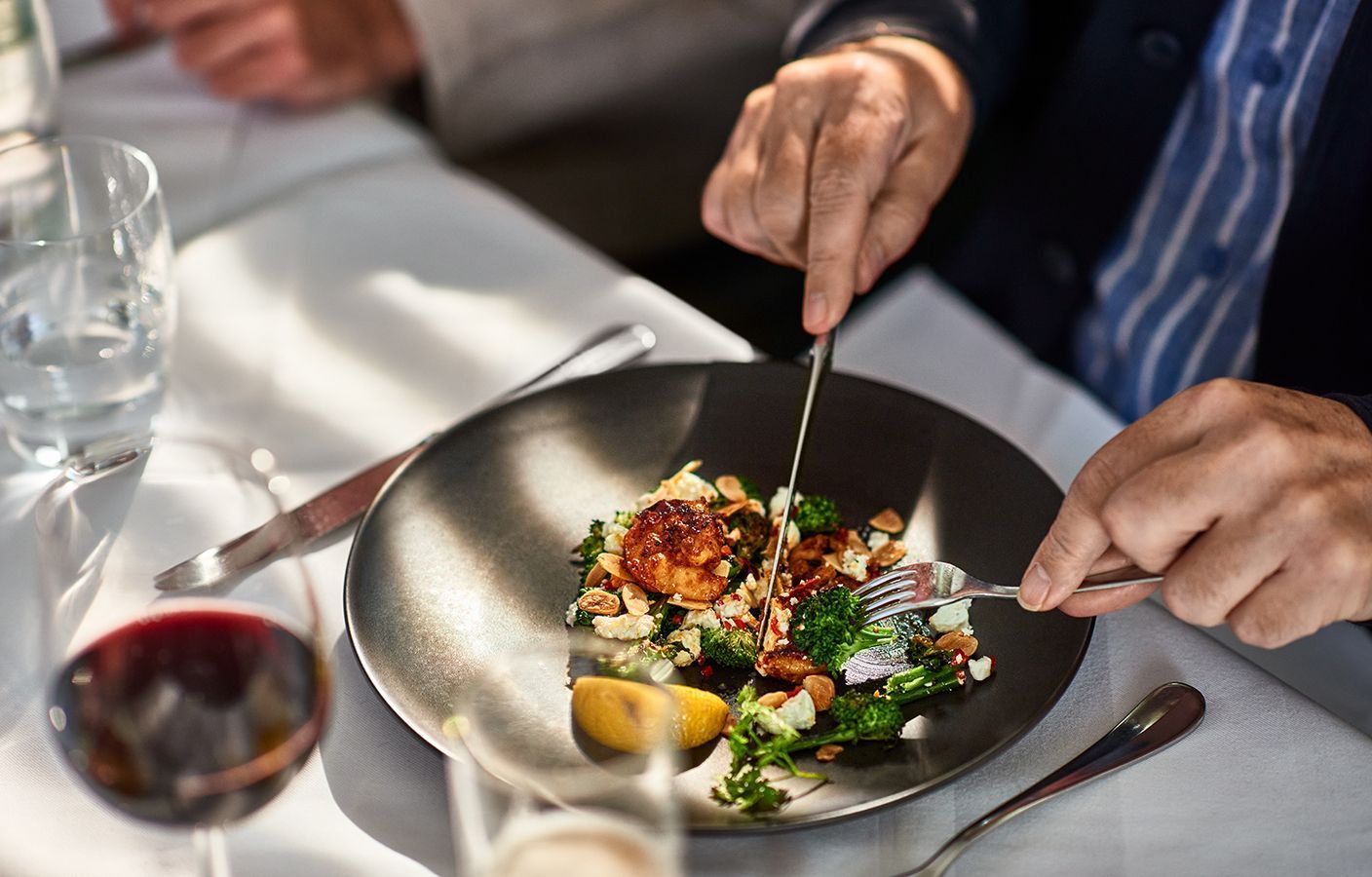 Persona che mangia l'insalata con coltello e forchetta a un tavolo; sono presenti un bicchiere di vino e un bicchiere d'acqua.
