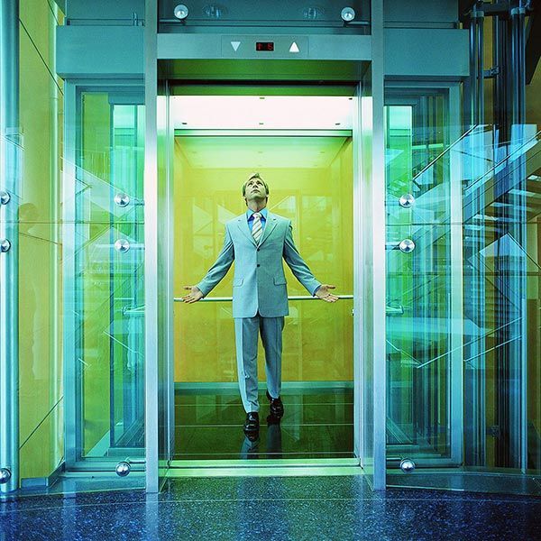 Businessman standing in a glass elevator, arms outstretched, with a green-lit background.