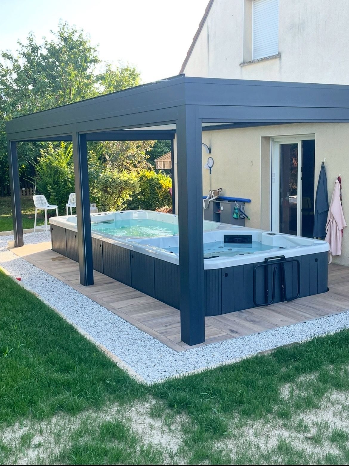 Un spa de nage rectangulaire sous une pergola gris foncé, à côté d'une maison avec un revêtement beige et une porte coulissante en verre.