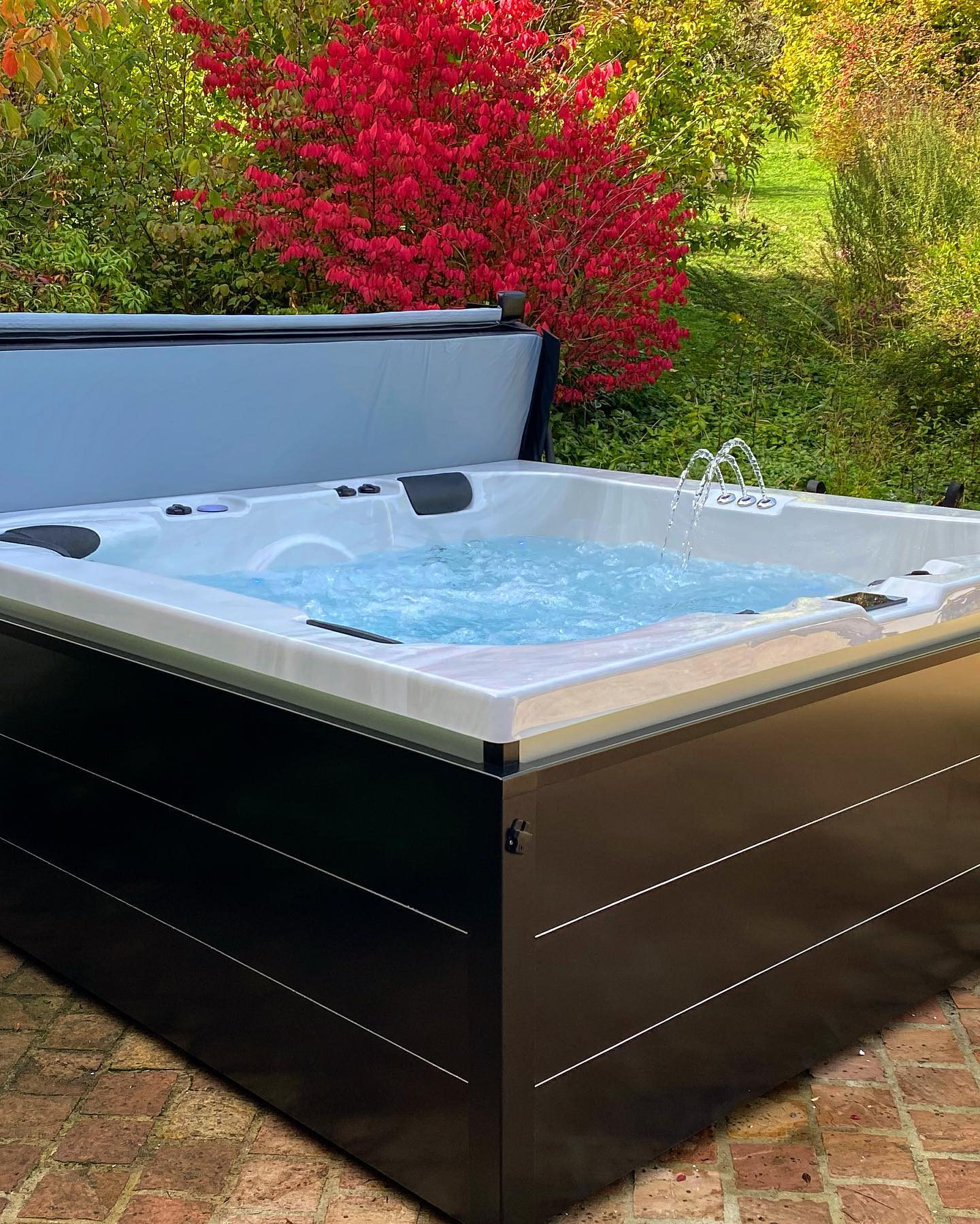 Un bain à remous est posé sur un patio en briques à côté d'un buisson aux feuilles rouges.