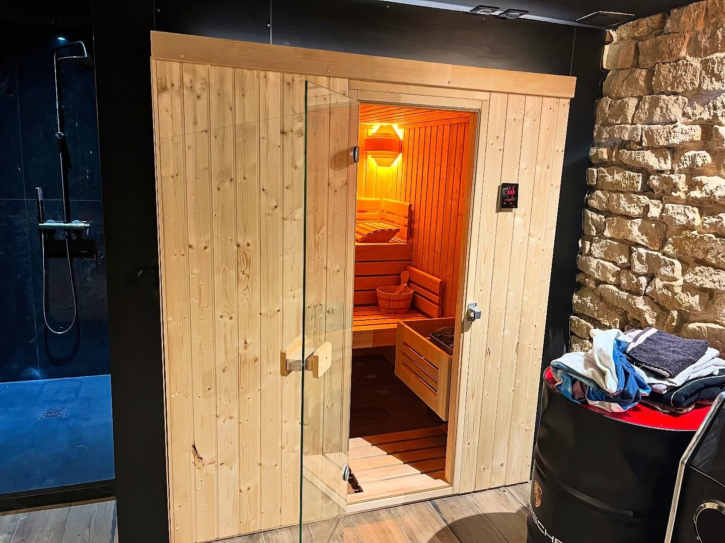Un sauna domestique en bois avec une porte vitrée, installé dans une pièce aux murs noirs et un mur en briques rustiques.