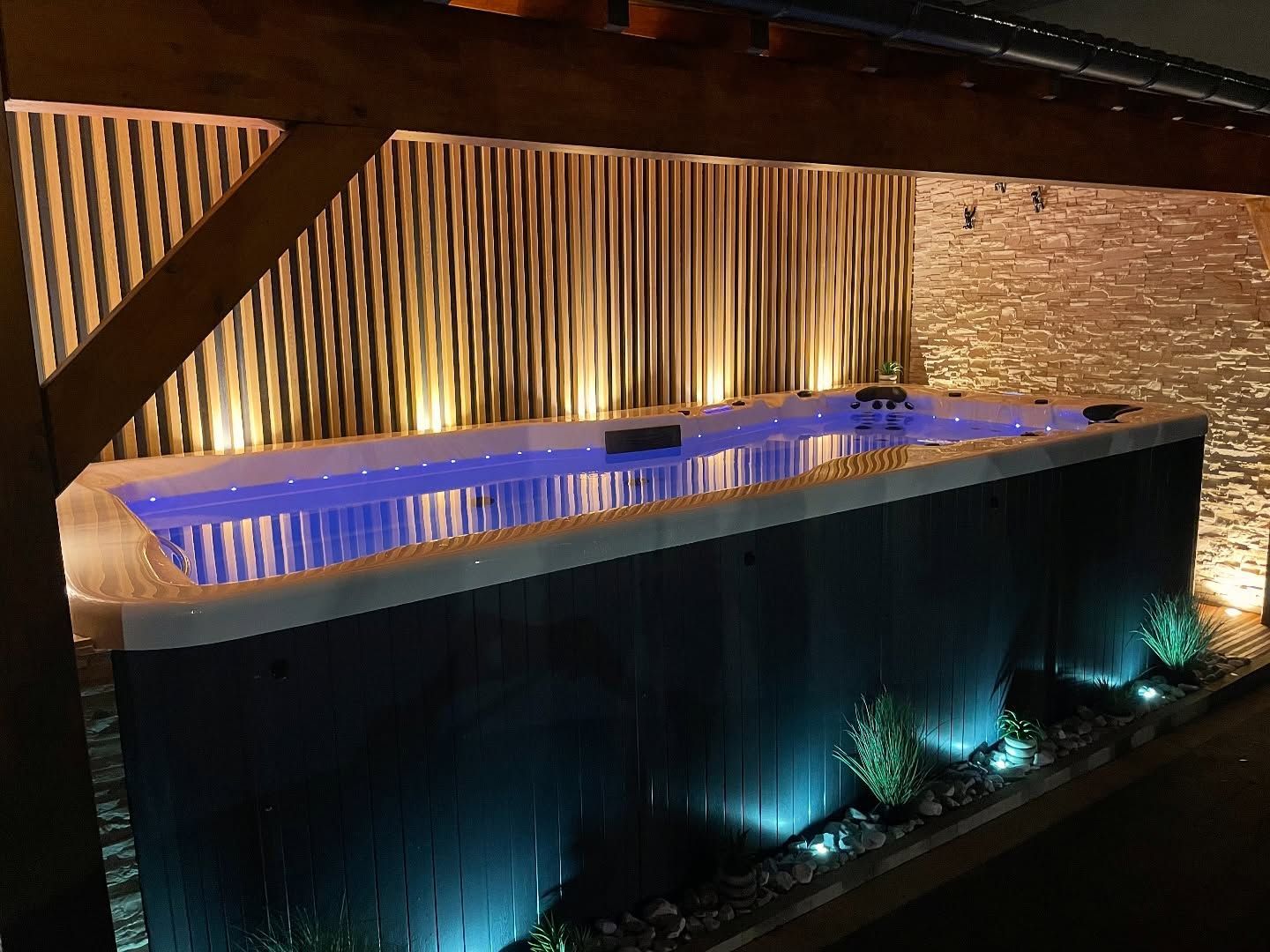 Un spa de nage rectangulaire aux eaux d'un bleu éclatant est installé sous une structure en bois, contre un mur texturé, le tout baigné d'un éclairage chaleureux.