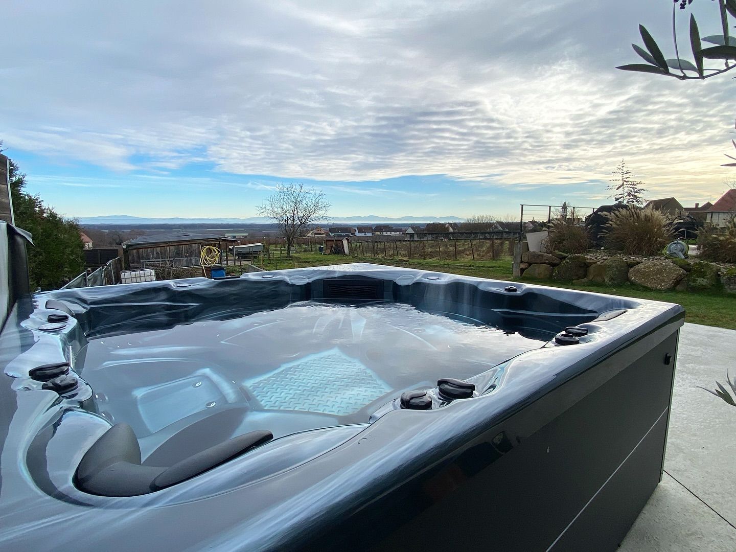 Un jacuzzi gris foncé rempli d'eau trône sur une terrasse surplombant un paysage champêtre sous un ciel nuageux.