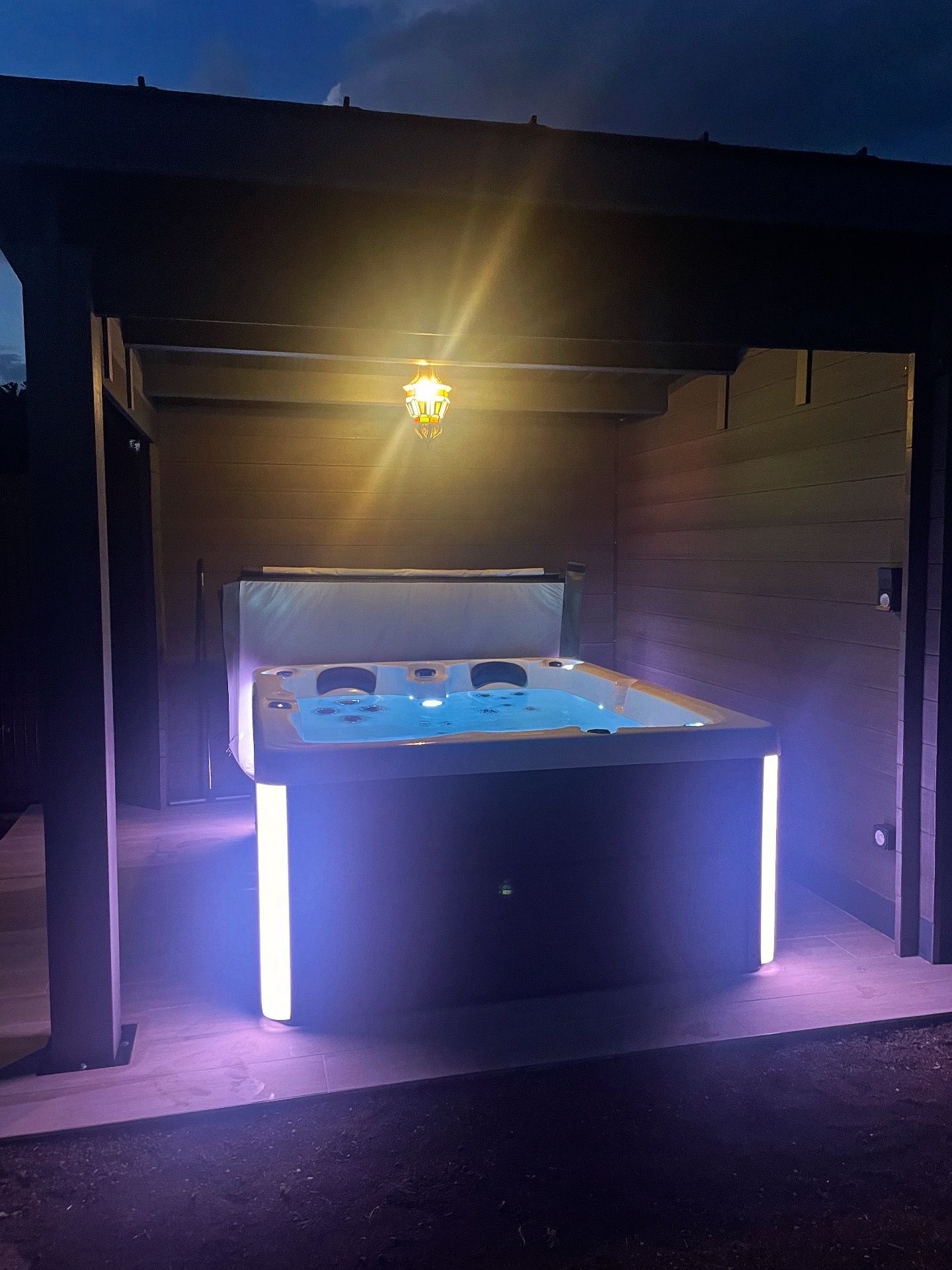 Un bain à remous éclairé avec des lumières violettes, sous une pergola en bois au crépuscule.