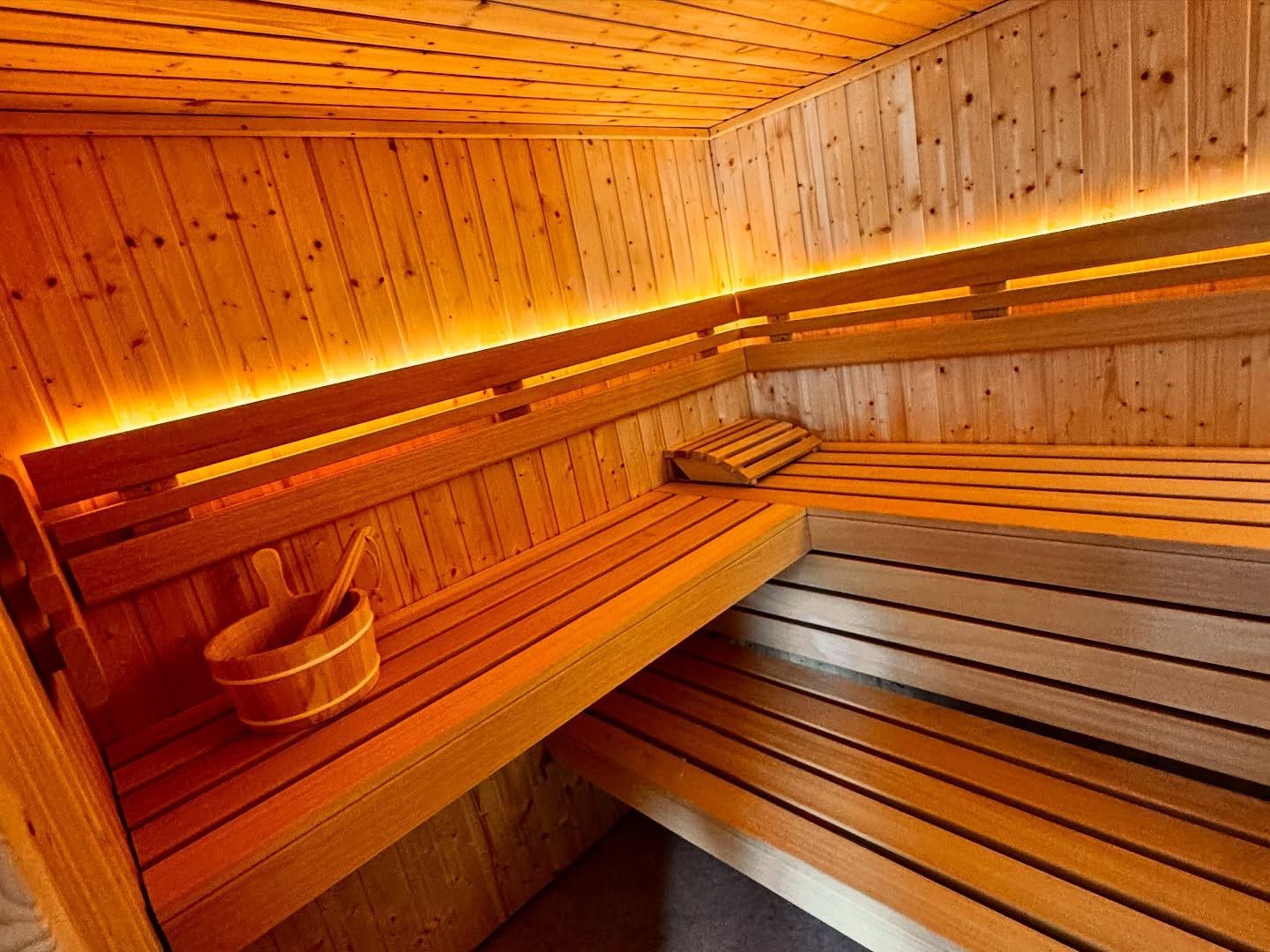 Une salle de sauna vide, aux murs en bois clair et aux banquettes étagées, éclairée par un éclairage LED indirect et chaleureux.