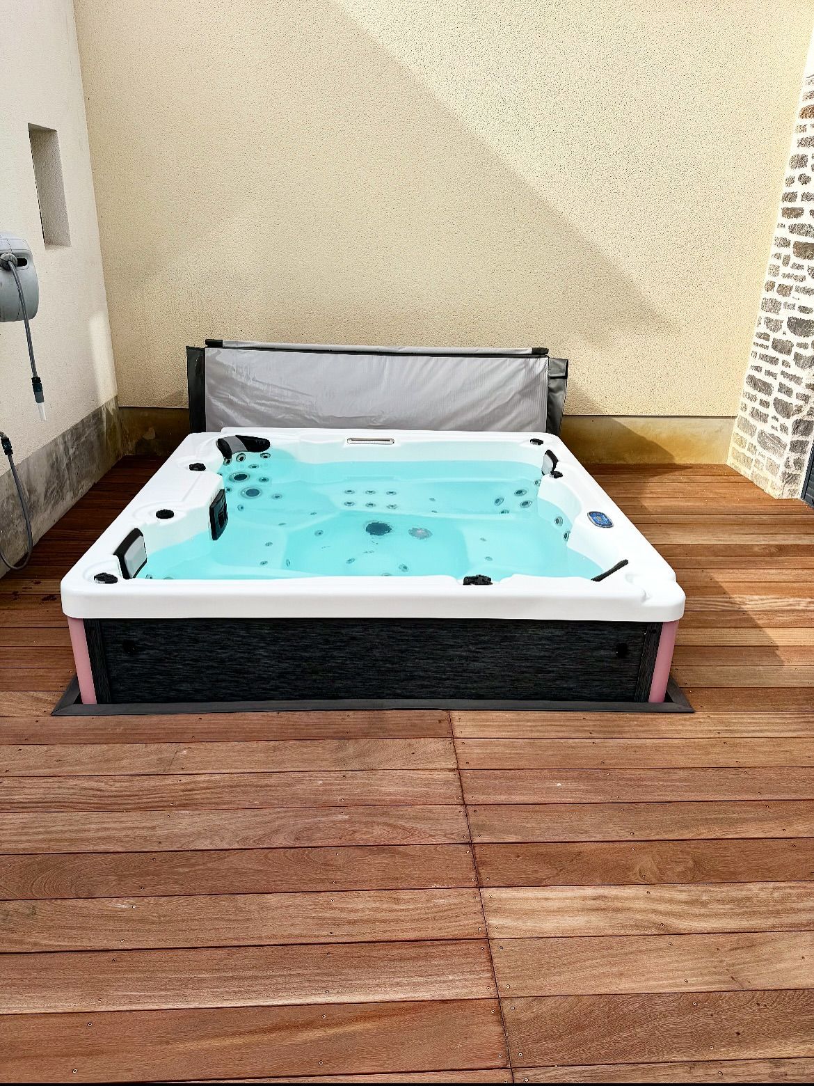 Un jacuzzi rectangulaire à l'eau turquoise, installé sur une terrasse en bois contre un mur texturé beige.