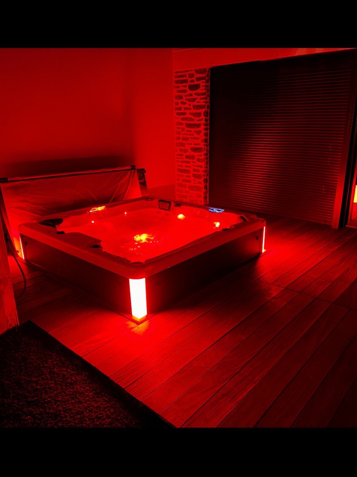 Un jacuzzi rectangulaire éclairé d'une lumière rouge éclatante, installé sur une terrasse en bois dans une pièce sombre aux murs texturés.