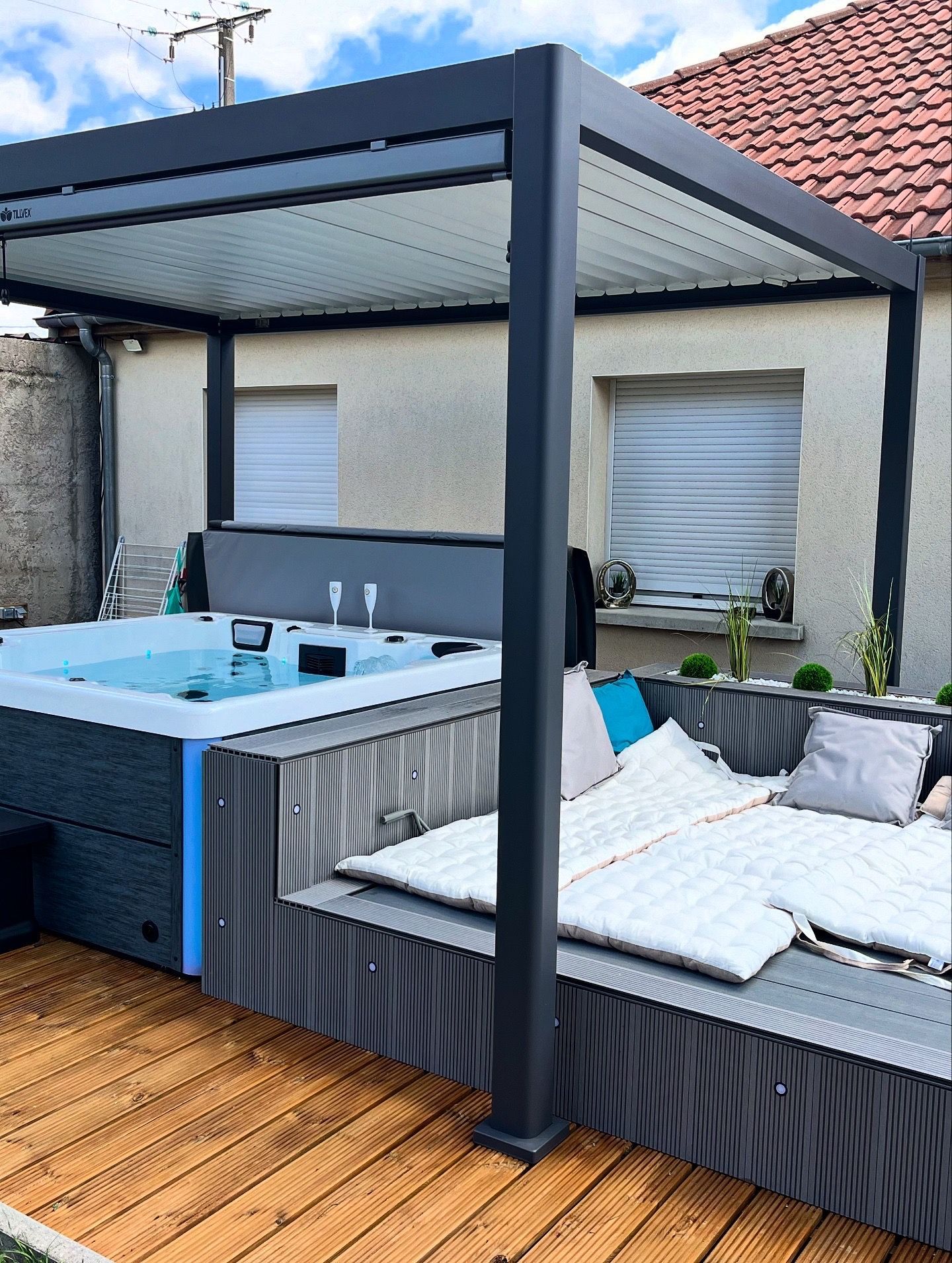 Jacuzzi et coin salon sous une pergola sur une terrasse en bois. Couleurs gris, bleu et blanc.