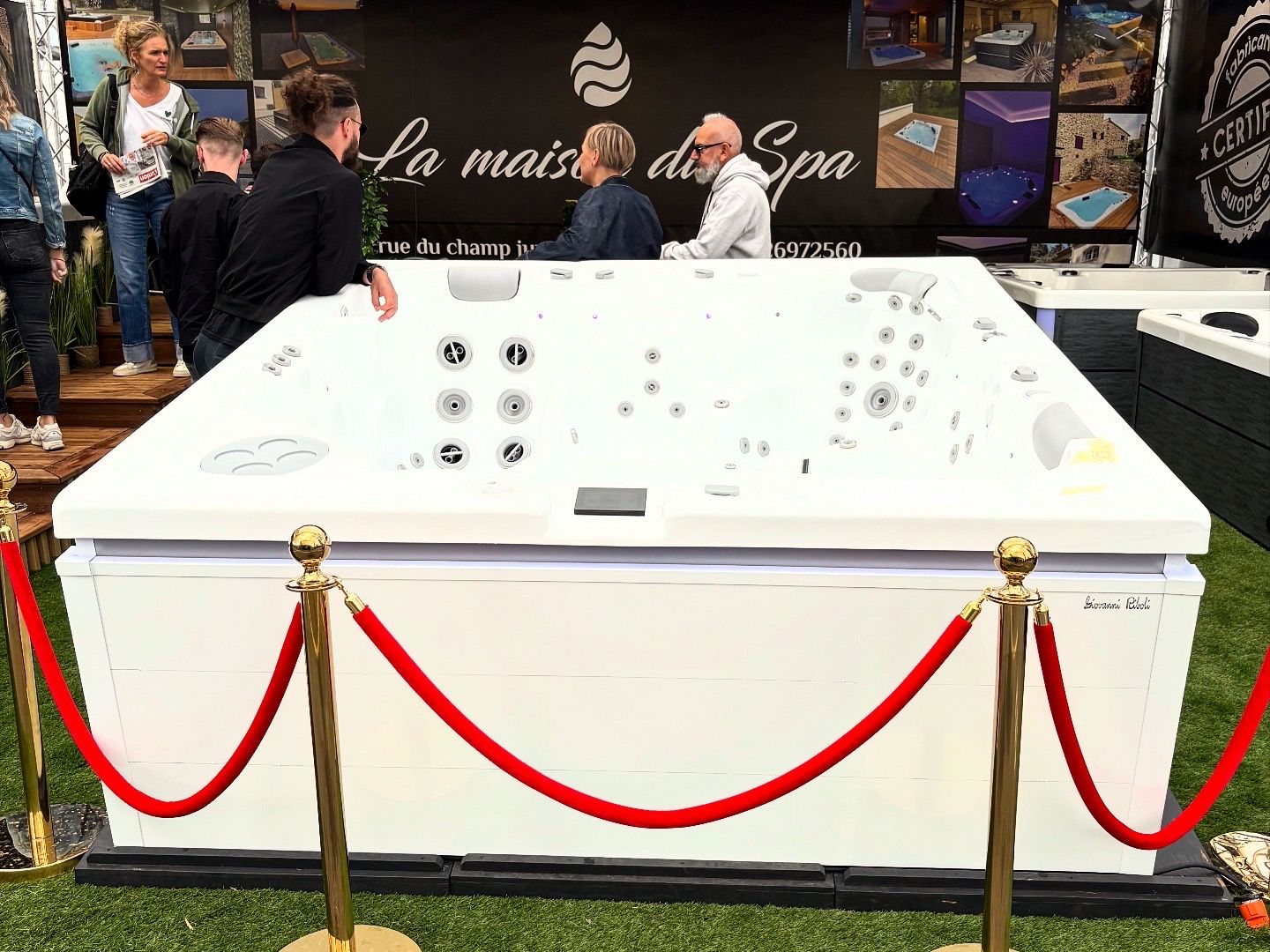 Un jacuzzi blanc avec une barrière en corde de velours rouge lors d'un salon professionnel, les gens le regardent.