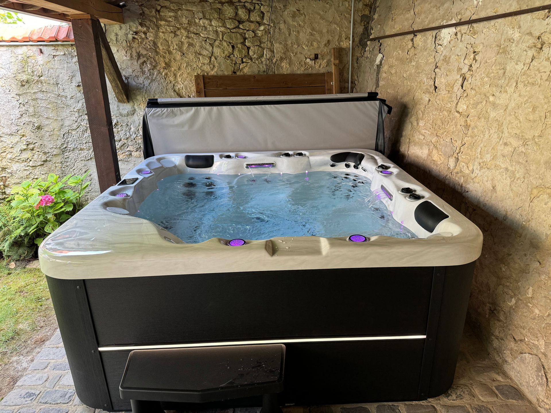Un jacuzzi noir et blanc est installé sous un patio couvert à côté d'un mur de pierre.