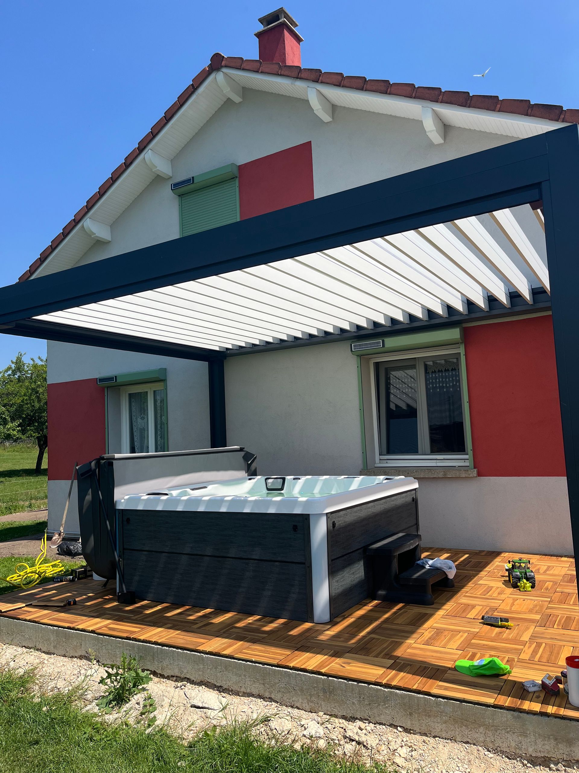 Un jacuzzi est installé sous une pergola devant une maison.