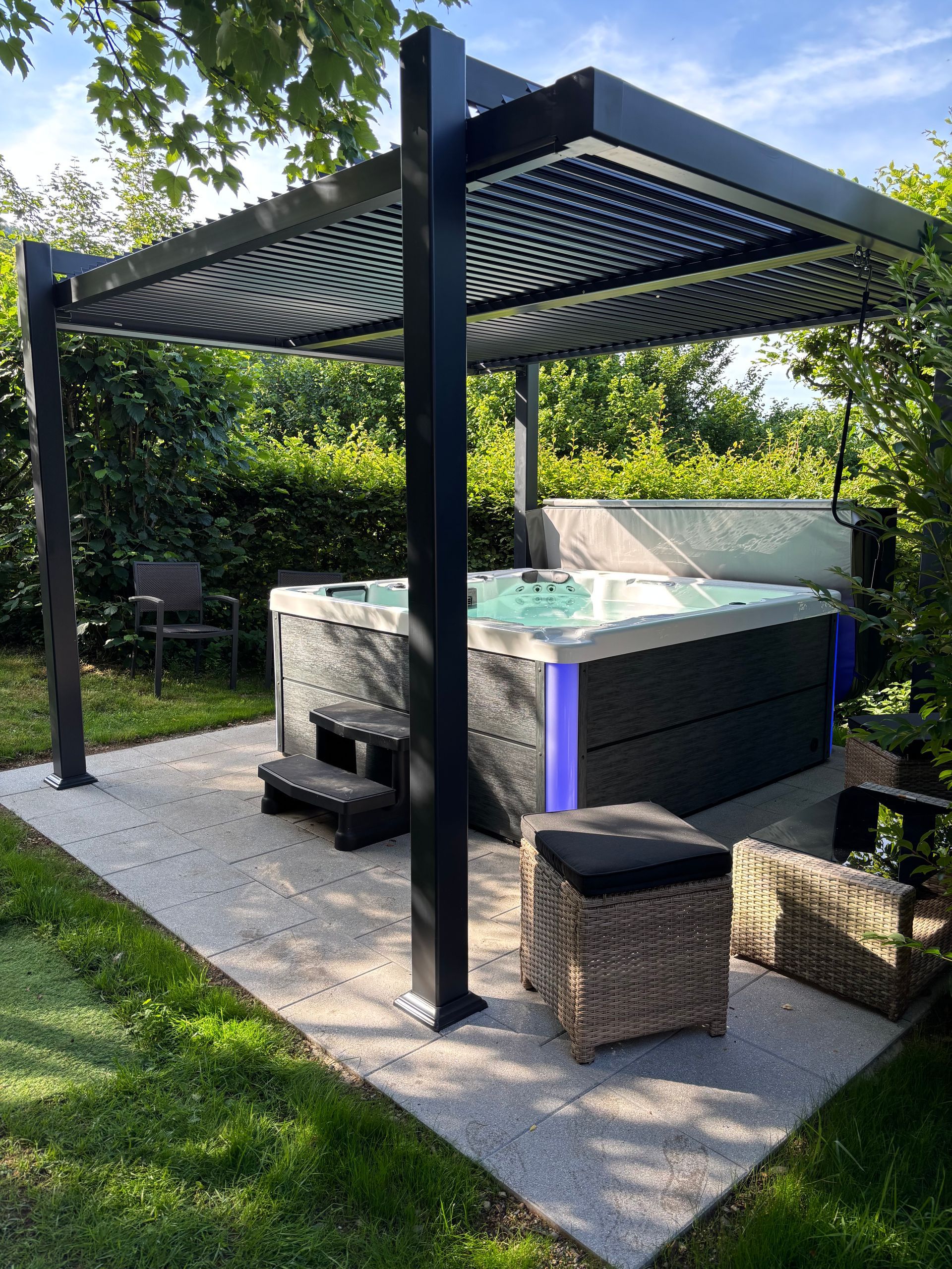 Spa sous pergola noire sur un patio en béton, avec marches, entouré de verdure.