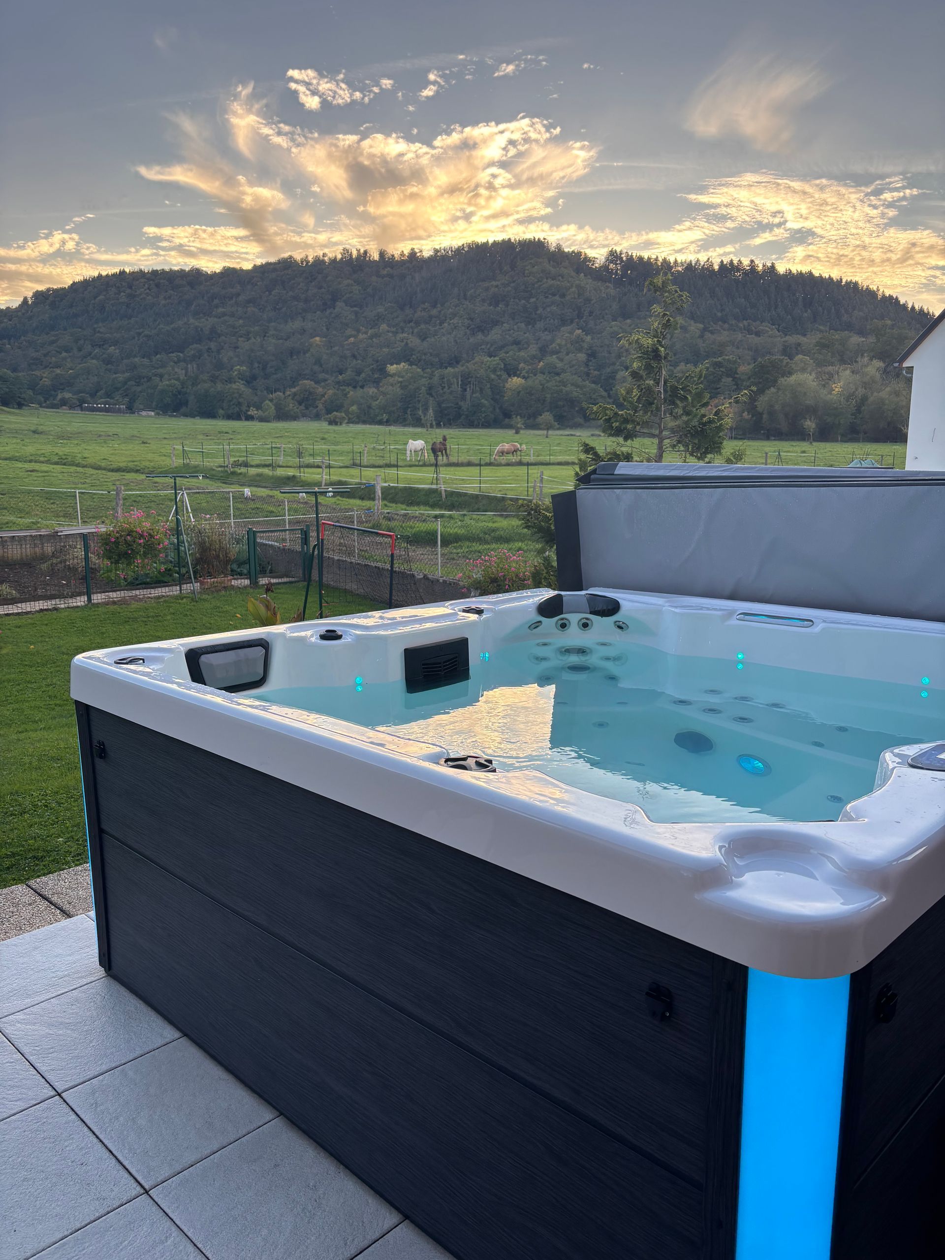 Bain à remous avec lumières bleues, avec vue sur un champ vert et une montagne au coucher du soleil.