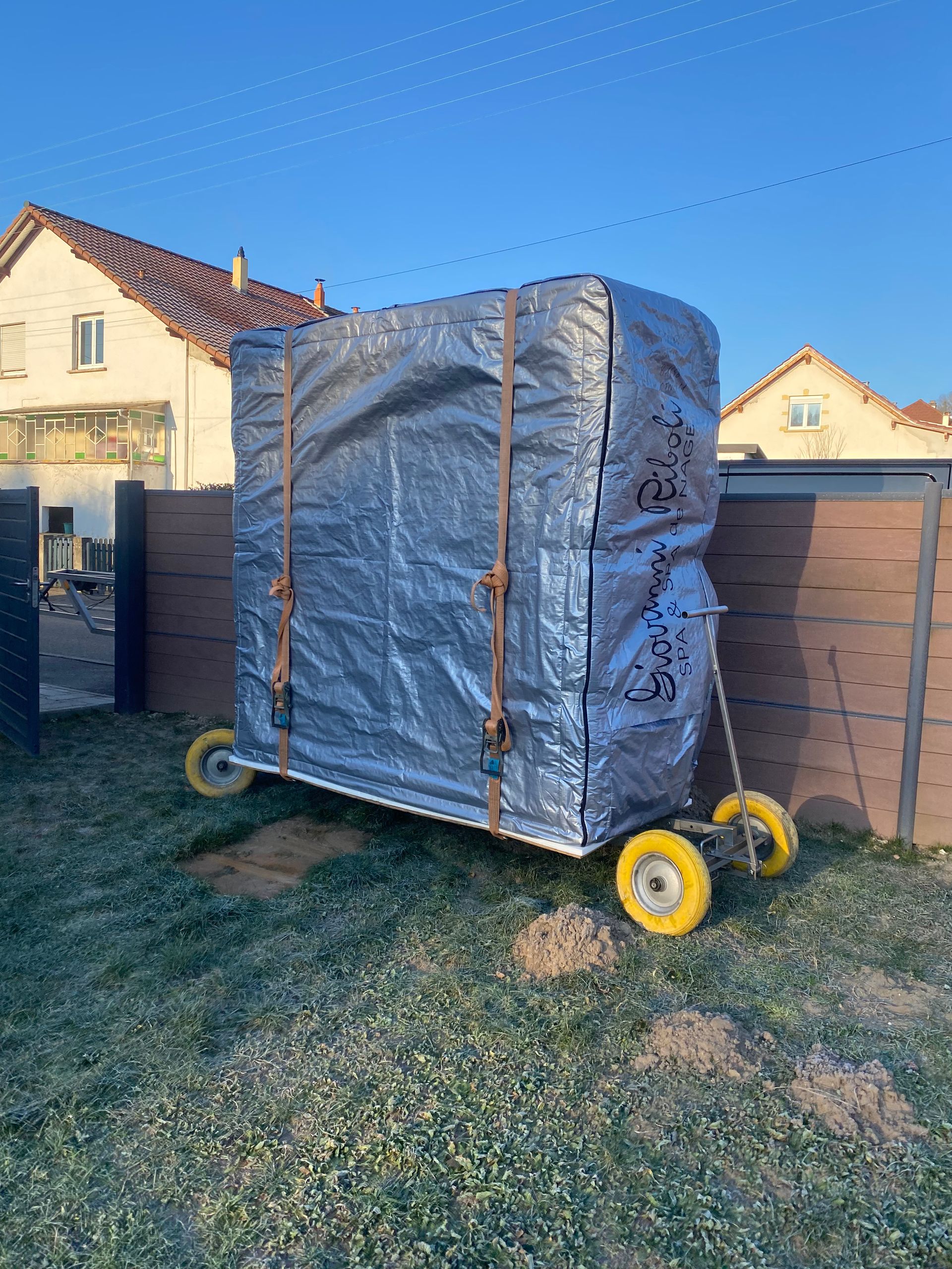 Une grande boîte est emballée dans du plastique et posée sur un chariot dans un champ herbeux.