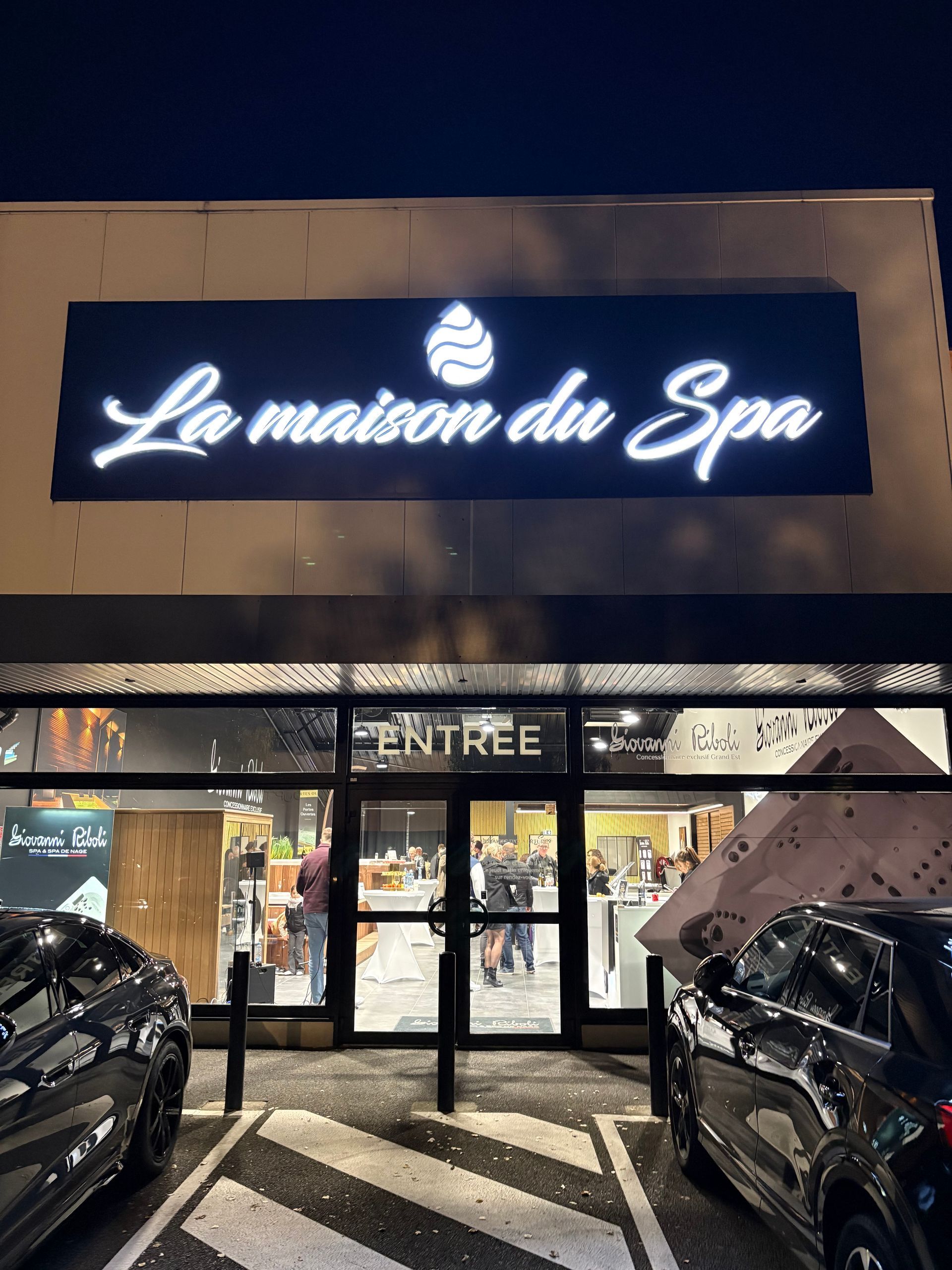 L'enseigne « La Maison du Spa » est illuminée la nuit. Le panneau « Entrée » est au-dessus de la porte. Voitures garées à proximité.