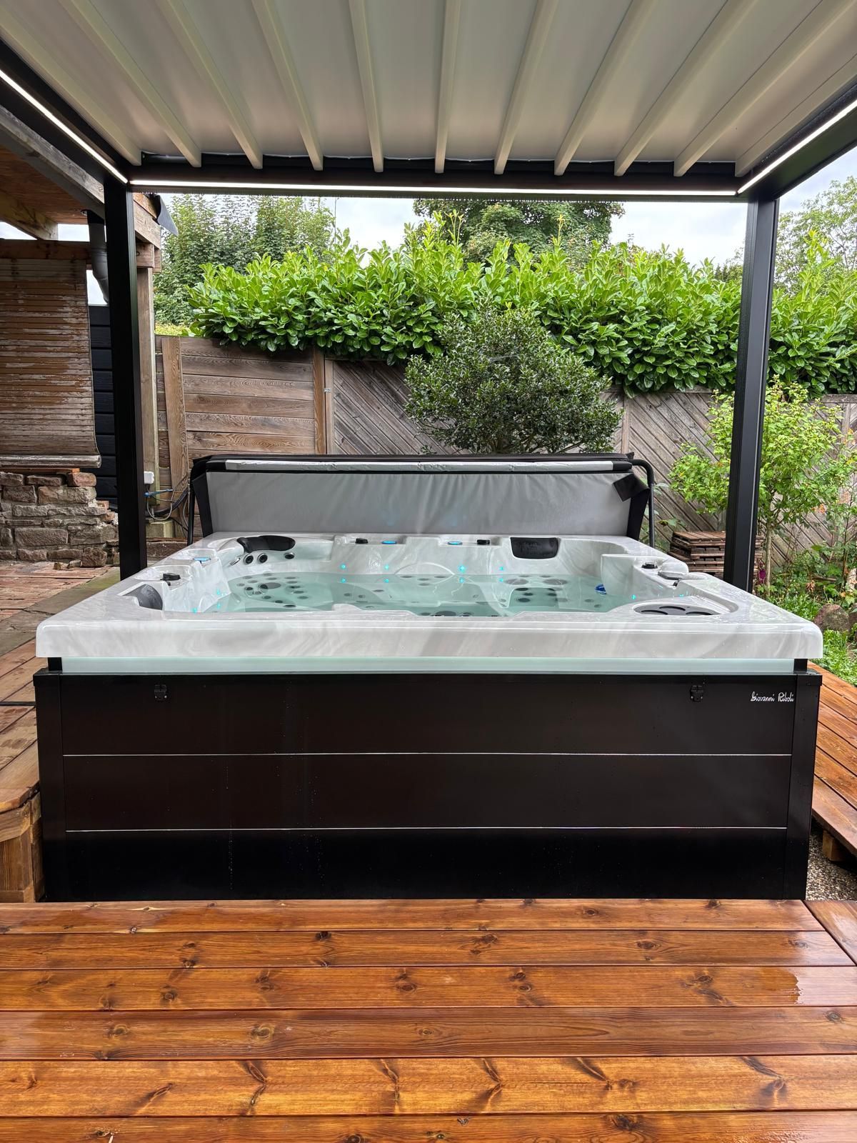 Un jacuzzi carré avec un intérieur en marbre clair et un revêtement extérieur brun foncé, installé sur une terrasse en bois sous un abri de patio en métal.