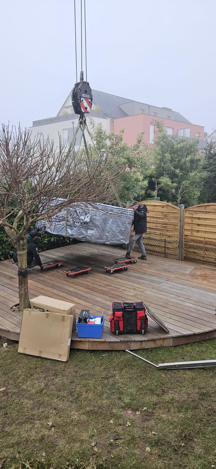 Une grue soulève un grand objet emballé sur une terrasse en bois. Deux personnes et des outils sont à proximité. Des maisons sont visibles à l'arrière-plan.