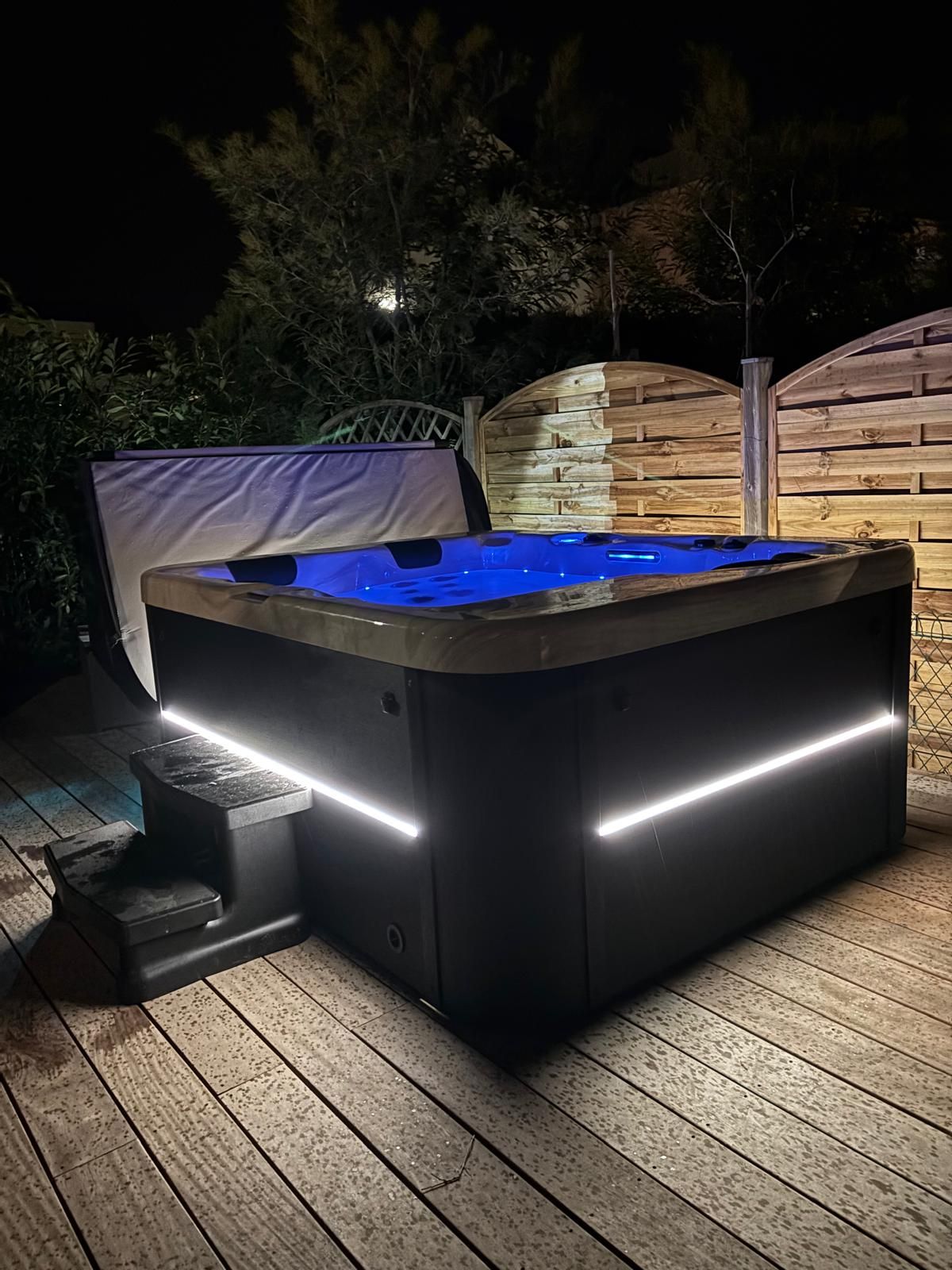 Bain à remous de nuit avec lumières intérieures bleues, bandes LED éclairées et terrasse en bois.