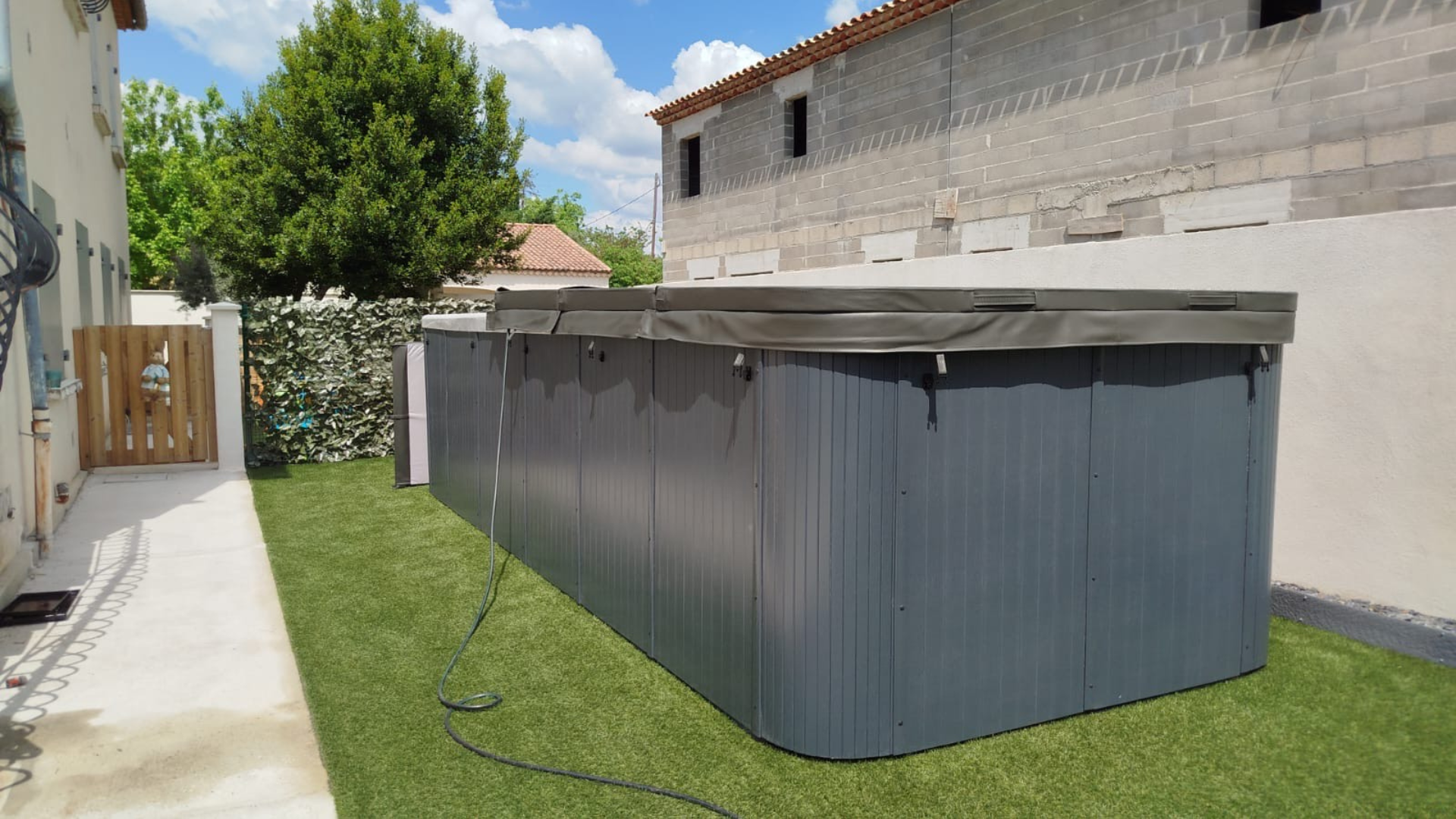 Un jacuzzi rectangulaire gris foncé avec un couvercle rigide est installé sur une parcelle de pelouse dans un jardin à côté d'une maison.