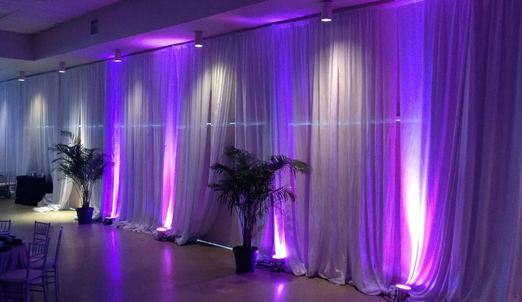 White drapes lit with purple uplighting in a venue, with potted plants.