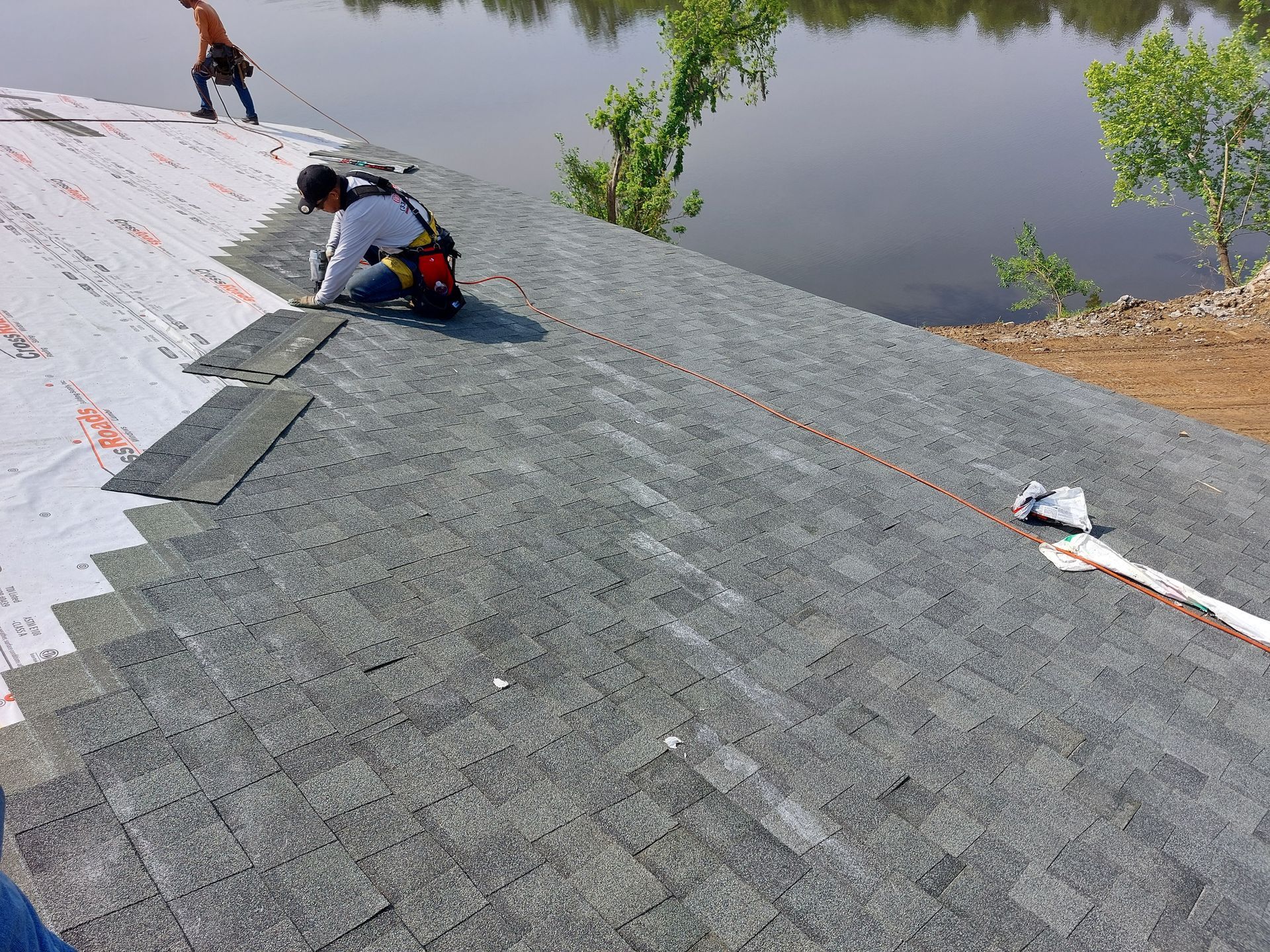 Workers laying gray roofing material on a sloped roof beside a body of water