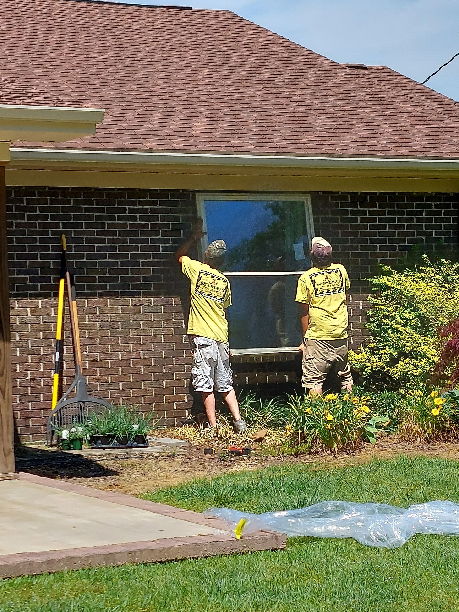 Two workers in yellow shirts install a window on a brick house exterior.