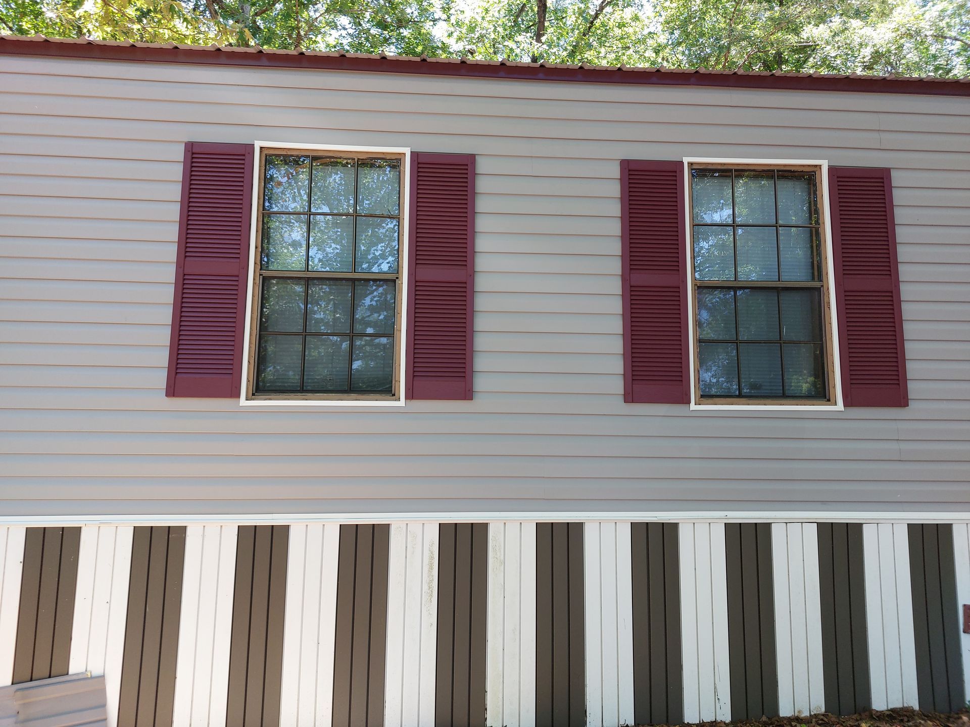 Mobile home exterior with two windows, maroon shutters, and a white-and-brown skirting panel